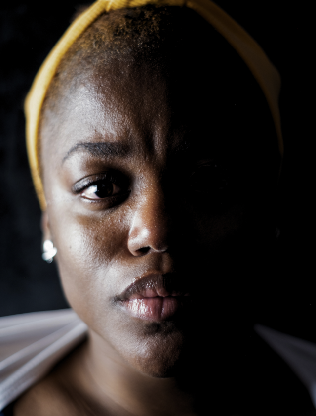 image of a human direclty facing the camera with half of their face lit and the other half in the dark, wearing a yellow headband and one diamond earring