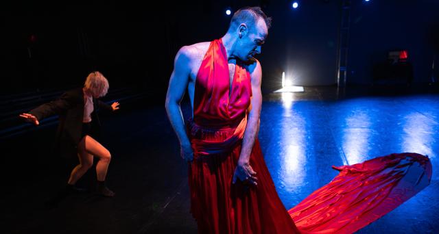 dancer standing and looking over left shoulder in red dress with dancer in background balancing with arms out and wearing a black coat