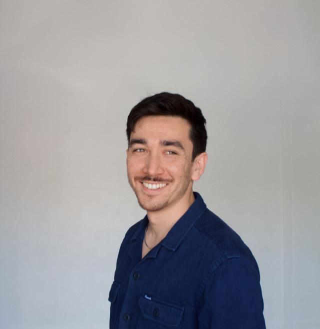 Person with dark hair and mustache wearing blue shirt against a grey backdrop