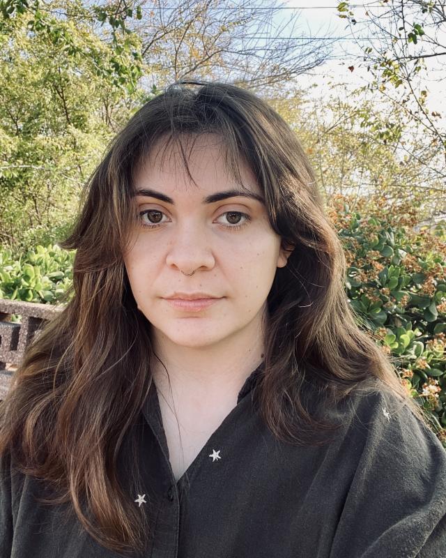 Person with dark brown hair and brown shirt in front of natural backdrop of trees and blue sky