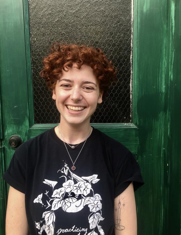 person with red curly hair and black t-shirt stands in front of a green door