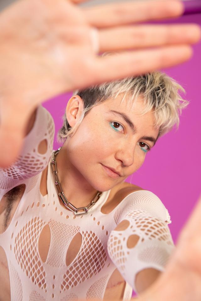 person with hands around camera lens framing their face with white top and standing in front of a pink background