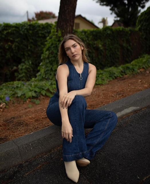 person with long brown hair sitting on log against natural backdrop wearing denim one piece