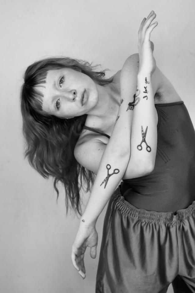 black and white photo of person crossing arms and leaning to the left with scissor tattoos on forearms and dark long hair