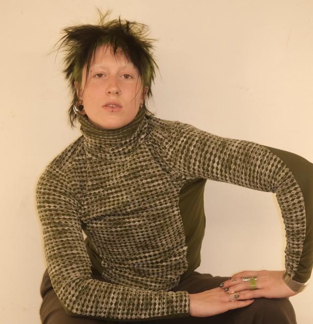 person with left arm bent on knee and crouching against a cream backdrop with a green turtleneck sweater and short hair