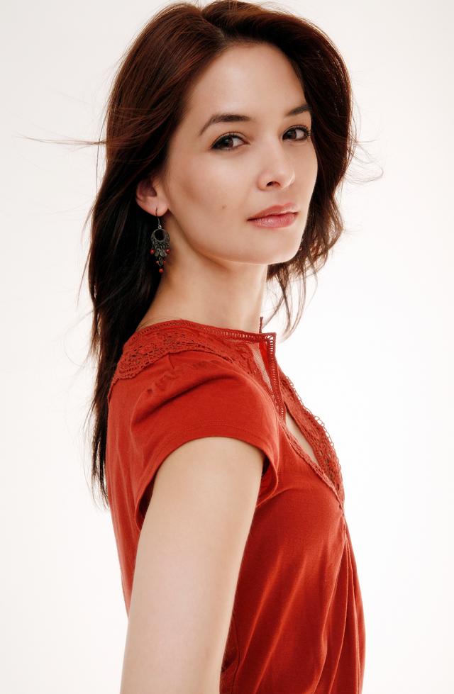 an image of a woman wearing a deep orange blouse against a white background. They are turned profile while their face is looking towards the camera