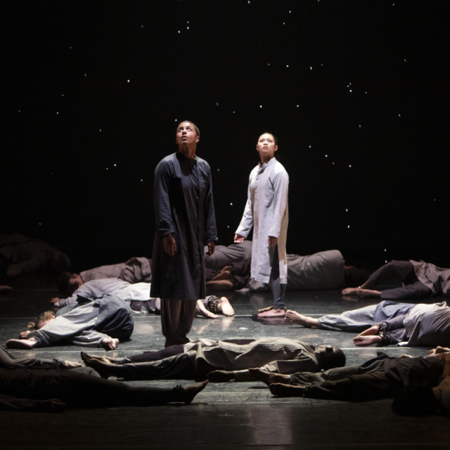 two people stand in black and white tunics looking at the sky with multiple people laying on the ground around their feet