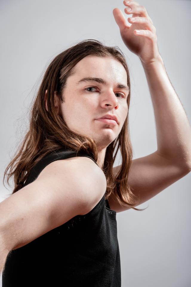 a photo of Will against a grey background with long brown hair positioned with their hand above their head and focused into the camera