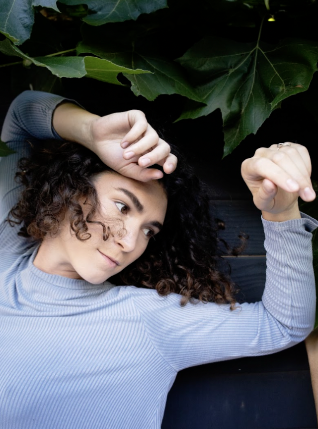 photo of a person with shoulder length curly brown hair, wearing a pale grey long sleeve with their arms overhead against green foliage