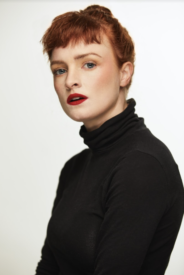 image of a person wearing all black and with read hair looking into the camera against a white background