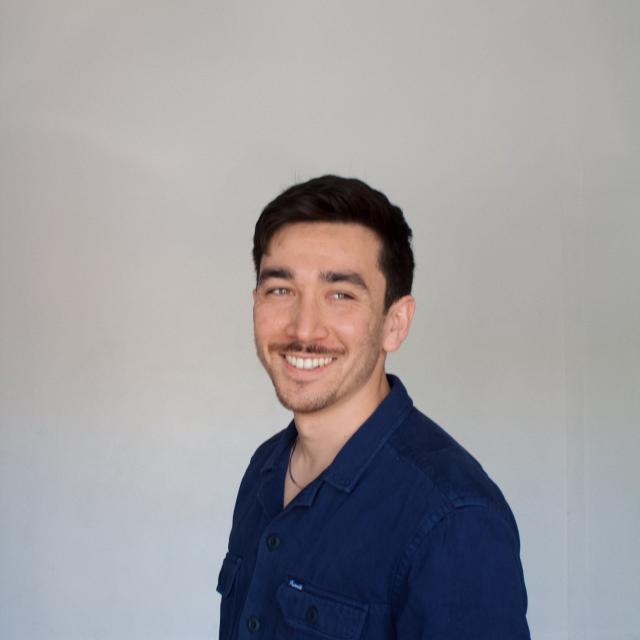 Person with dark hair and mustache wearing blue shirt against a grey backdrop