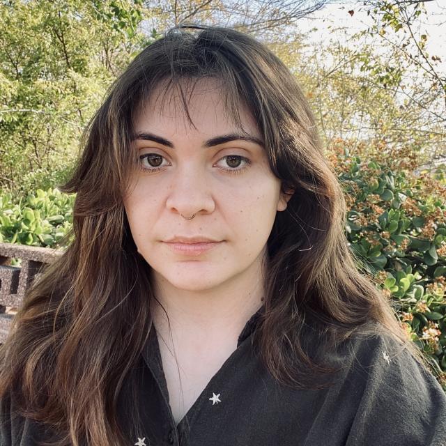 Person with dark brown hair and brown shirt in front of natural backdrop of trees and blue sky