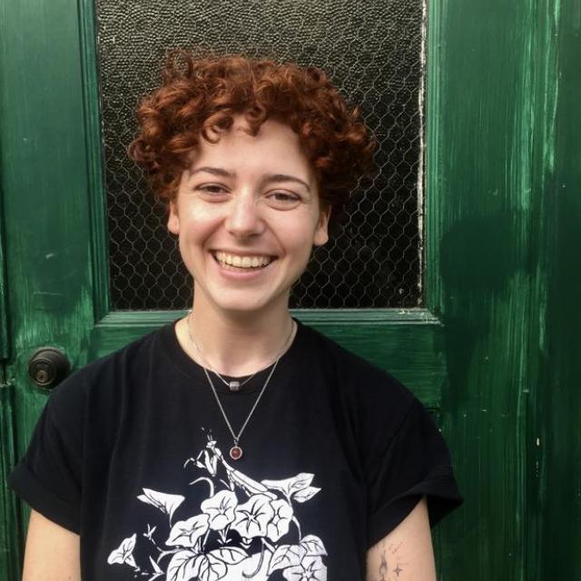 person with red curly hair and black t-shirt stands in front of a green door