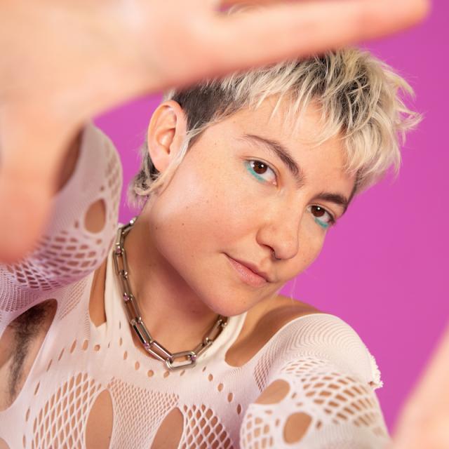 person with hands around camera lens framing their face with white top and standing in front of a pink background