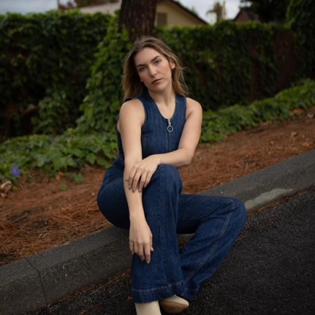 person with long brown hair sitting on log against natural backdrop wearing denim one piece