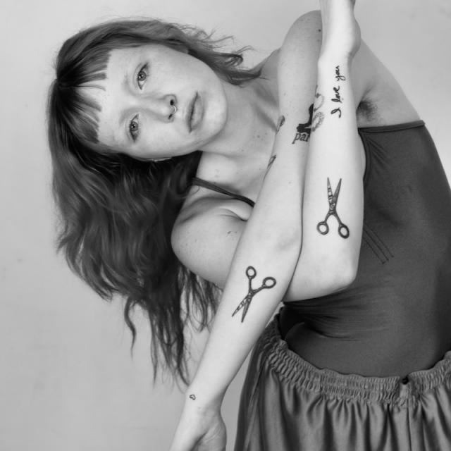 black and white photo of person crossing arms and leaning to the left with scissor tattoos on forearms and dark long hair