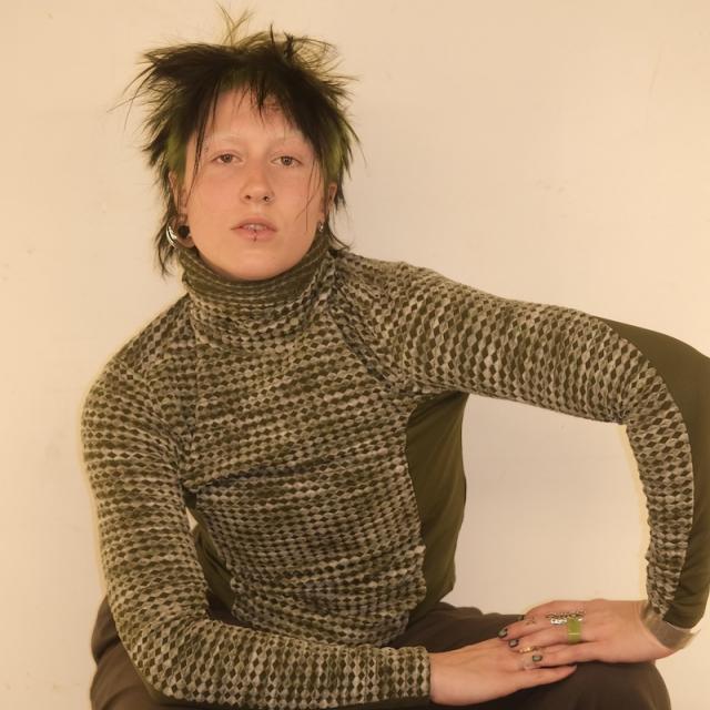 person with left arm bent on knee and crouching against a cream backdrop with a green turtleneck sweater and short hair