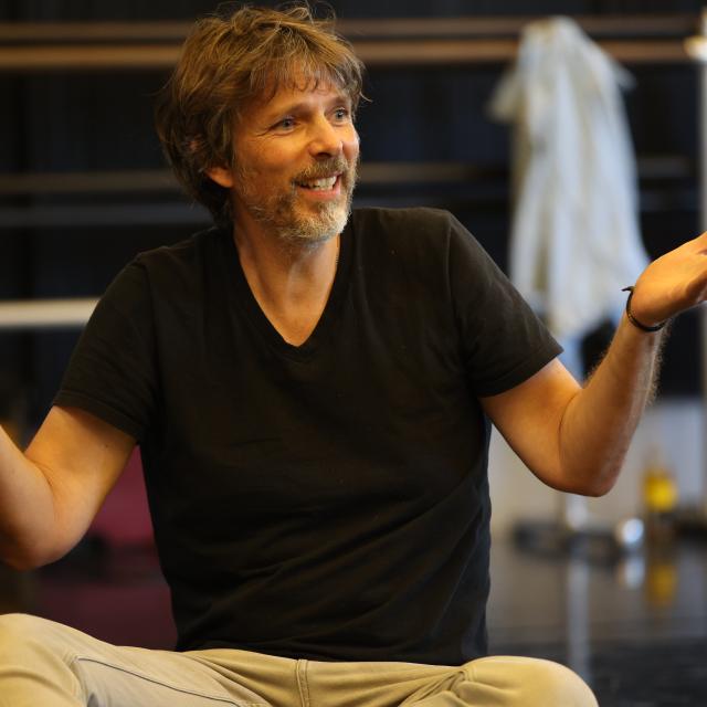 an image of a man with scruffy brown and grey facial hair sitting criss crossed with his hands open like they're holding trays