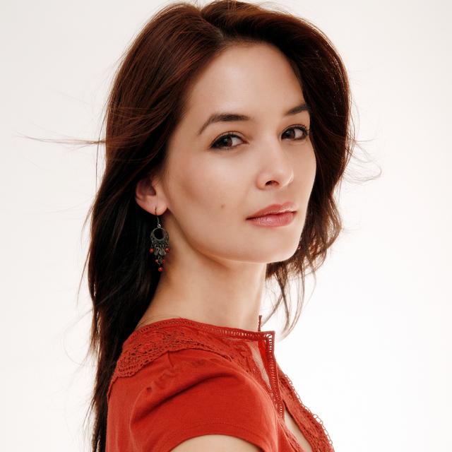 an image of a woman wearing a deep orange blouse against a white background. They are turned profile while their face is looking towards the camera