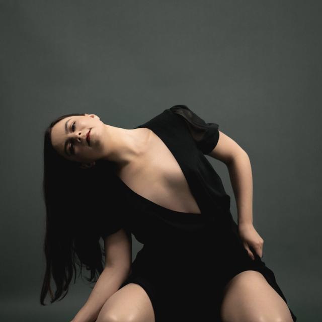 A photo of Anna Long against a dark grey backdrop. She has fair skin, and is wearing a black dress with a plunging neckline. She is placed down in a leaned over position cascading over her right ribs as her hair is draping down. Her focus is towards the camera with a bold red lip.