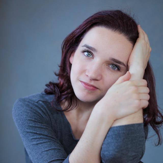 a photo of a person with shoulder length hair, and their hands caressing the side of their face wearing blue against a blue background