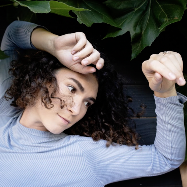 photo of a person with shoulder length curly brown hair, wearing a pale grey long sleeve with their arms overhead against green foliage