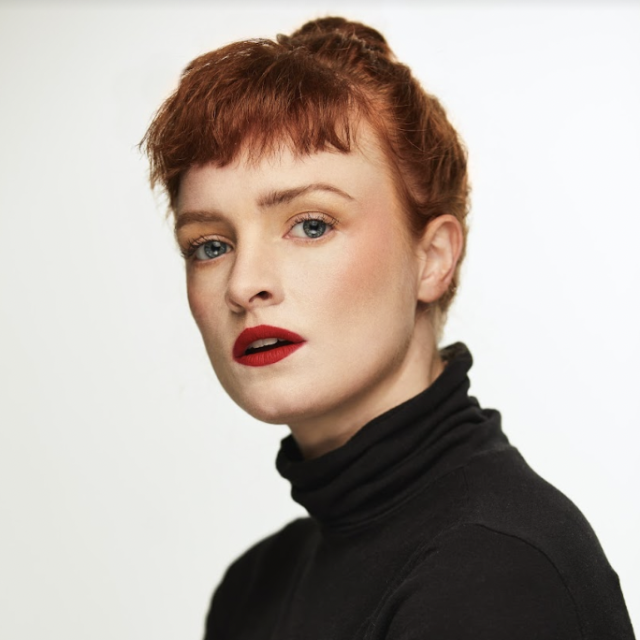 image of a person wearing all black and with read hair looking into the camera against a white background